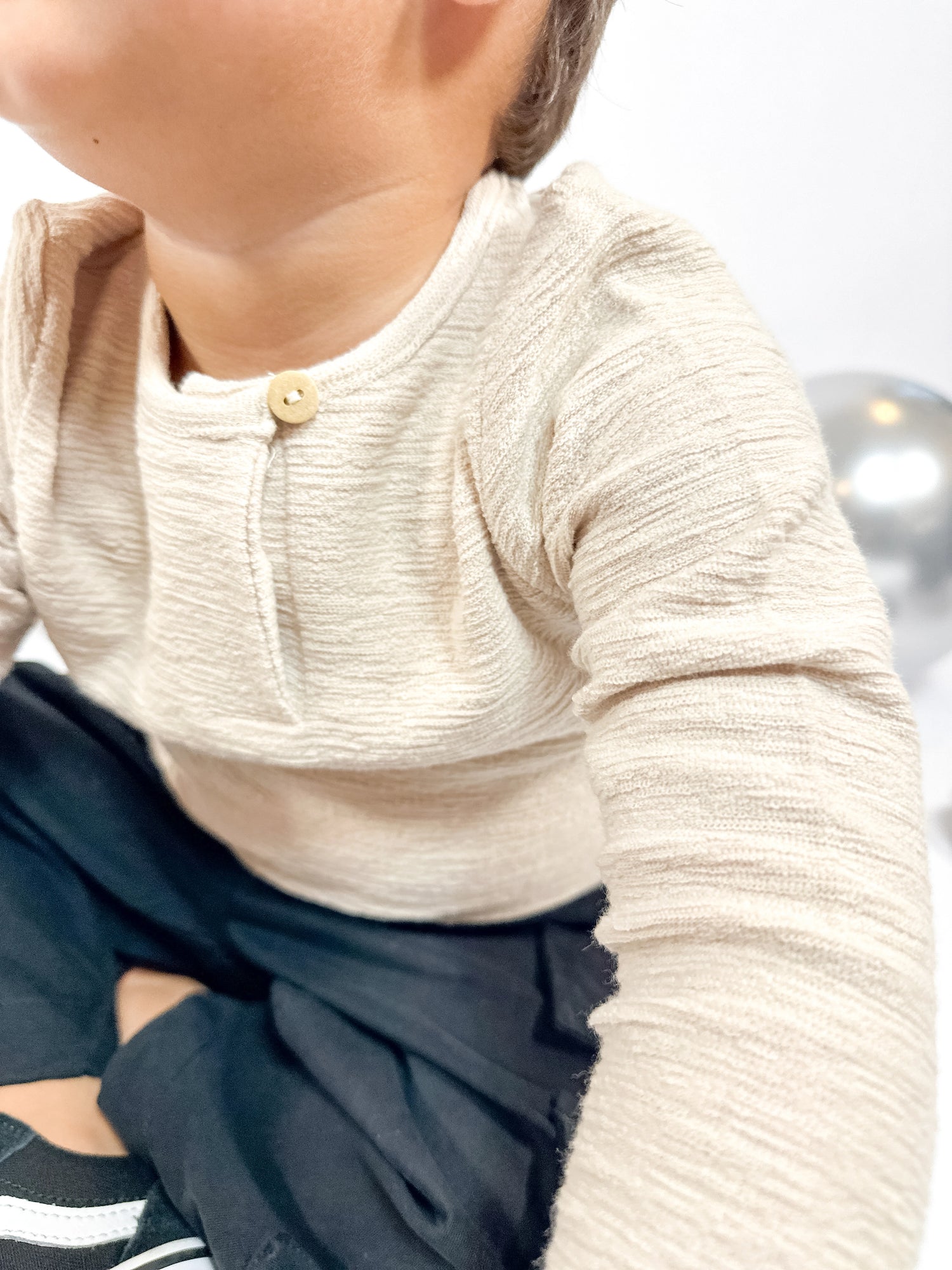 Close-up of little aurel model wearing a beige long-sleeve shirt with a button, sitting on a white surface.