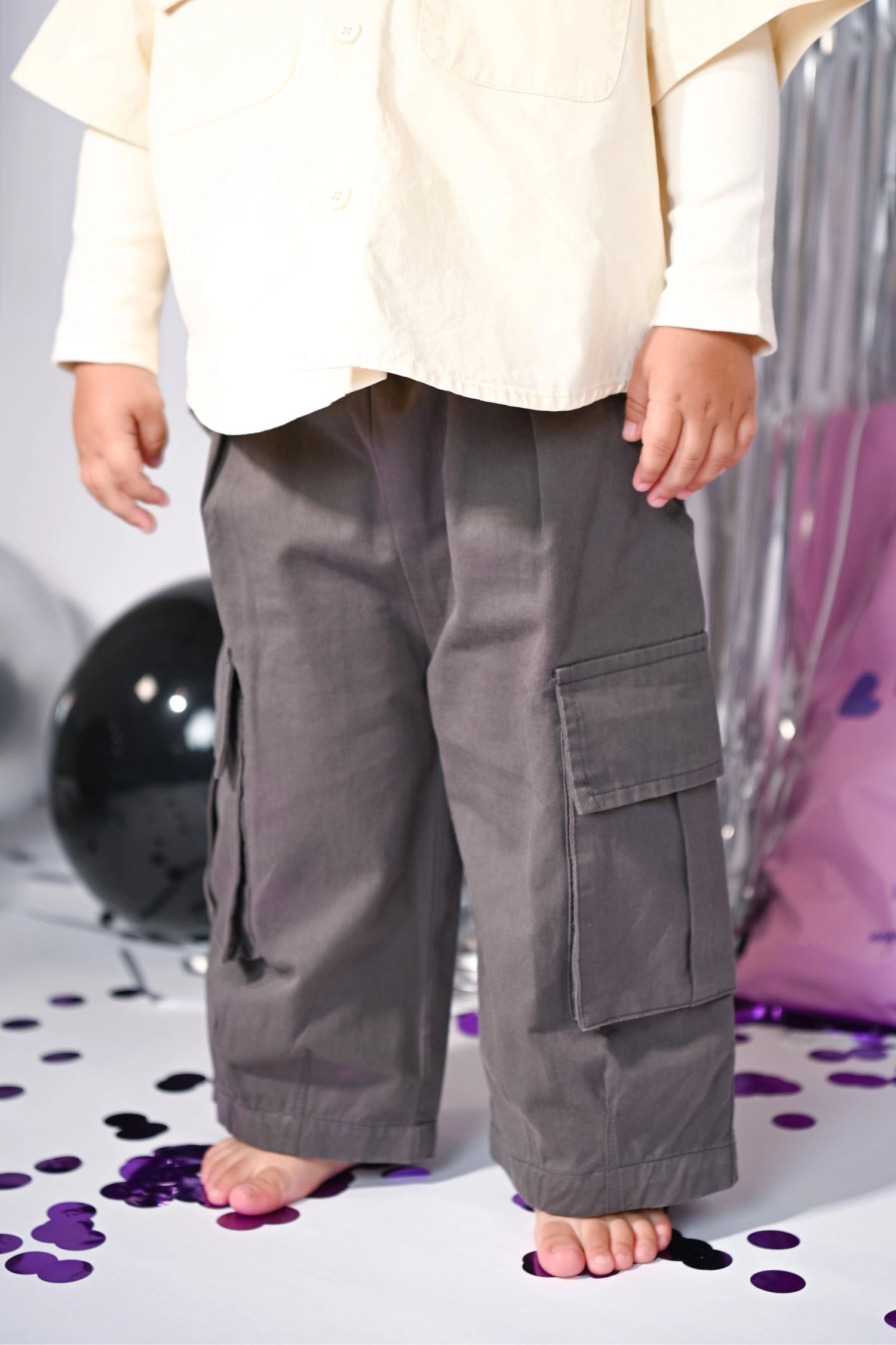 Little Aurel Model wearing a creamy yellow shirt with embroidered patches and gray cargo pants on a reflective surface.