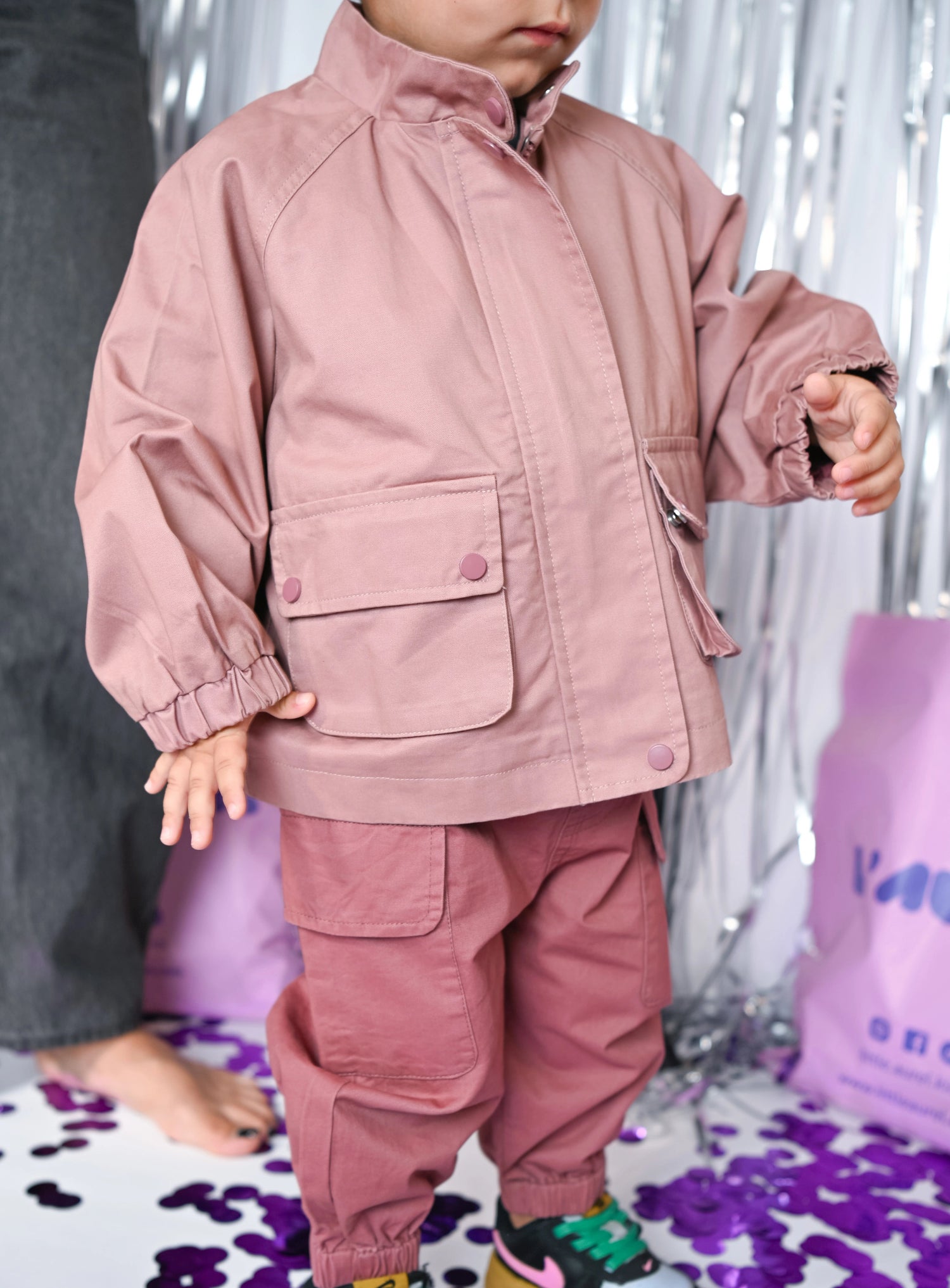 Child wearing a pink outfit by Little Aurel with colorful shoes on a patterned floor.