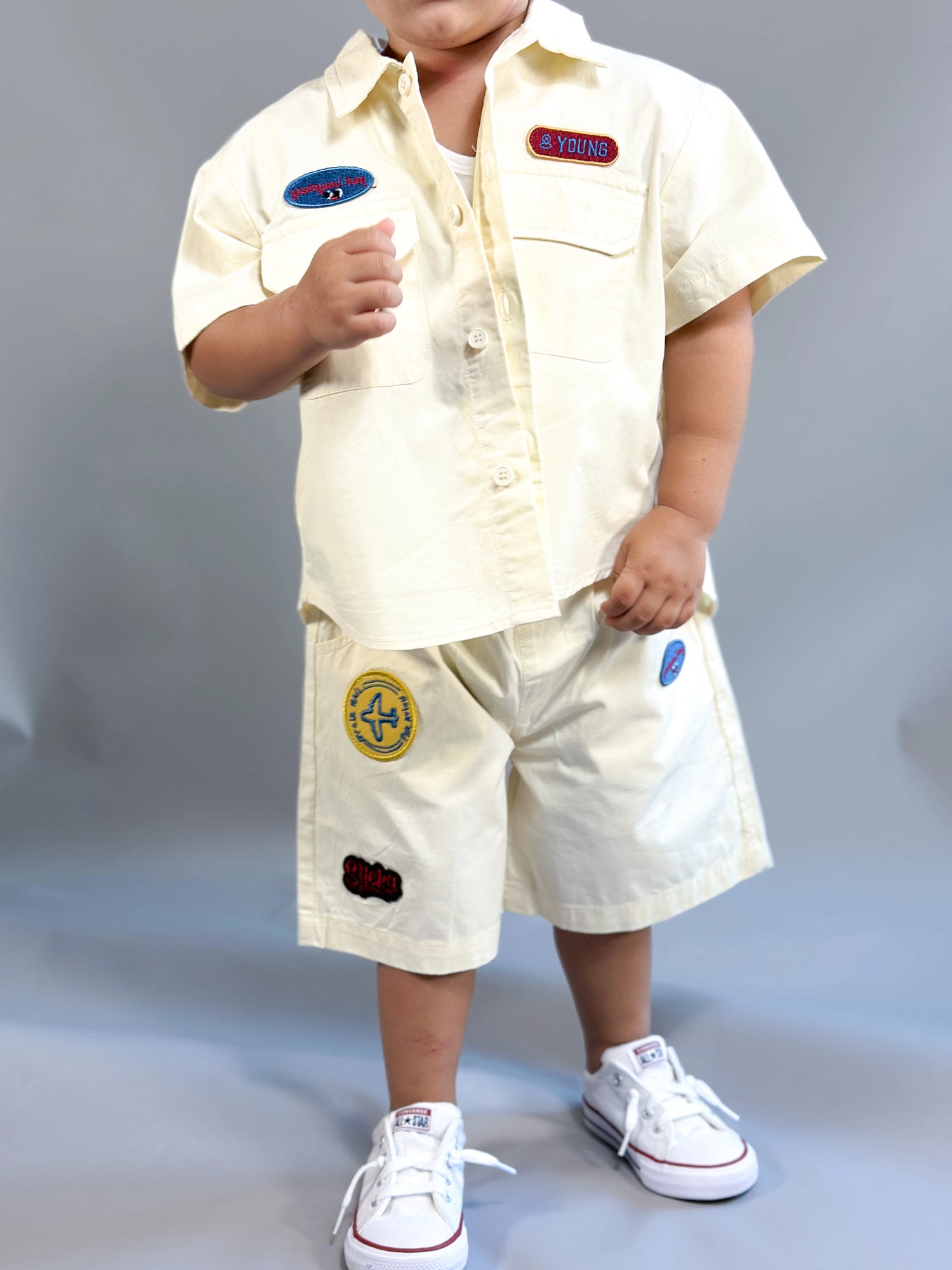Child wearing a cream-colored outfit with patches on a gray background by L'aurel