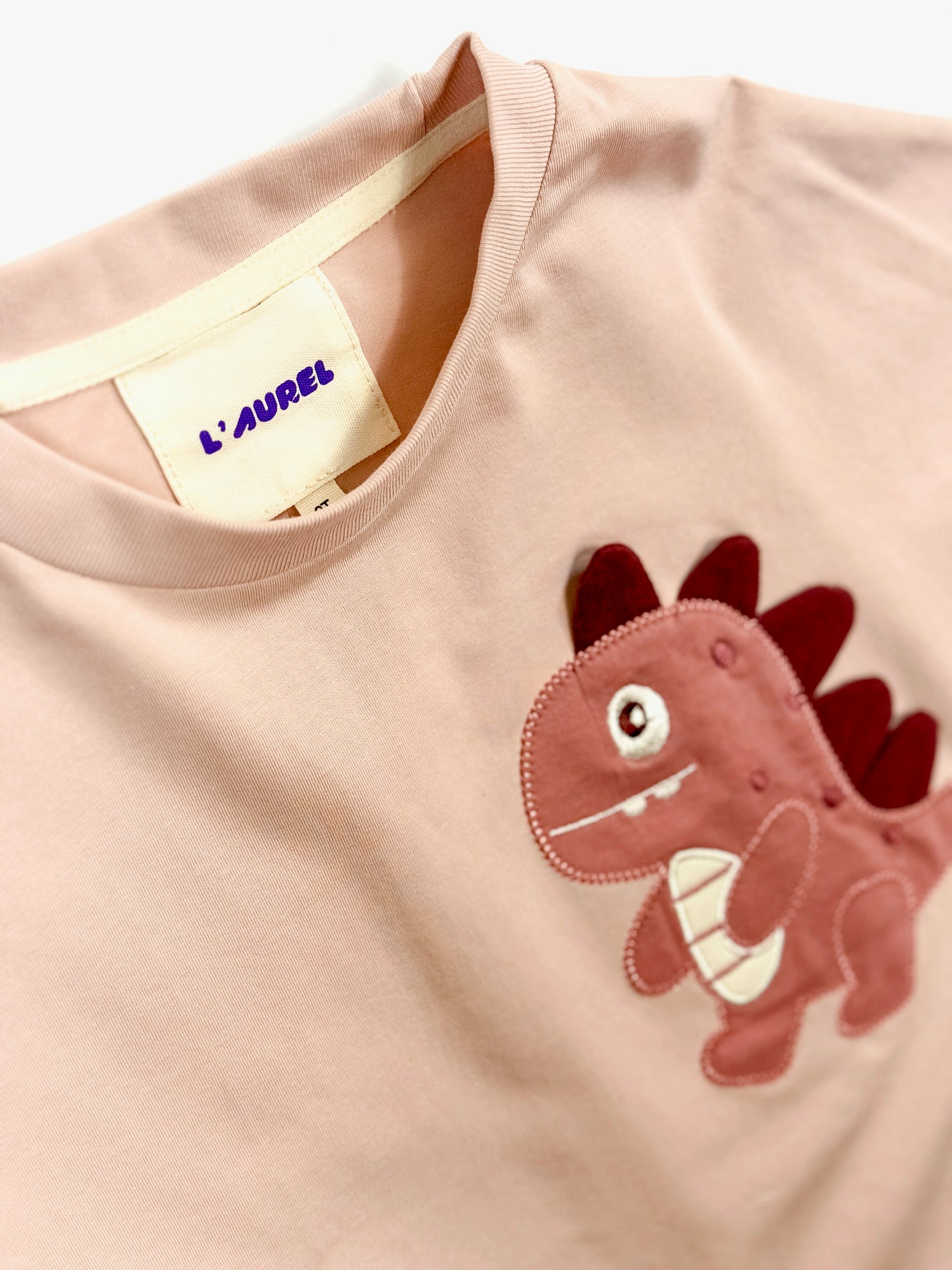 Pink shirt with a dinosaur applique and L'Aurel label on a white background
