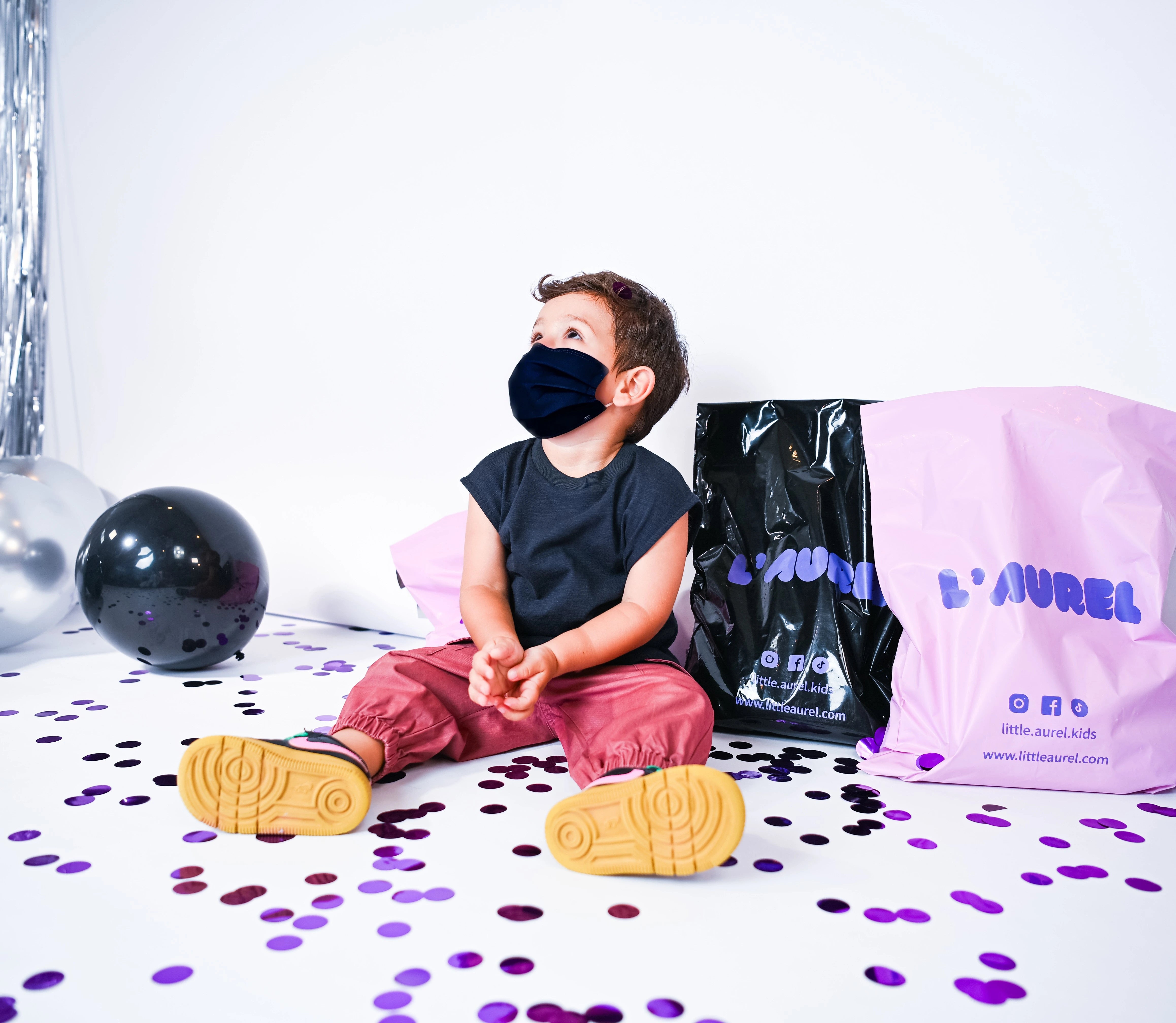 Child sitting on a white surface with confetti, wearing a black mask and colorful shoes, with a branded bag in the background.