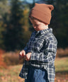 Child wearing a plaid shirt and brown beanie outdoors with trees in the background for Little Aurel