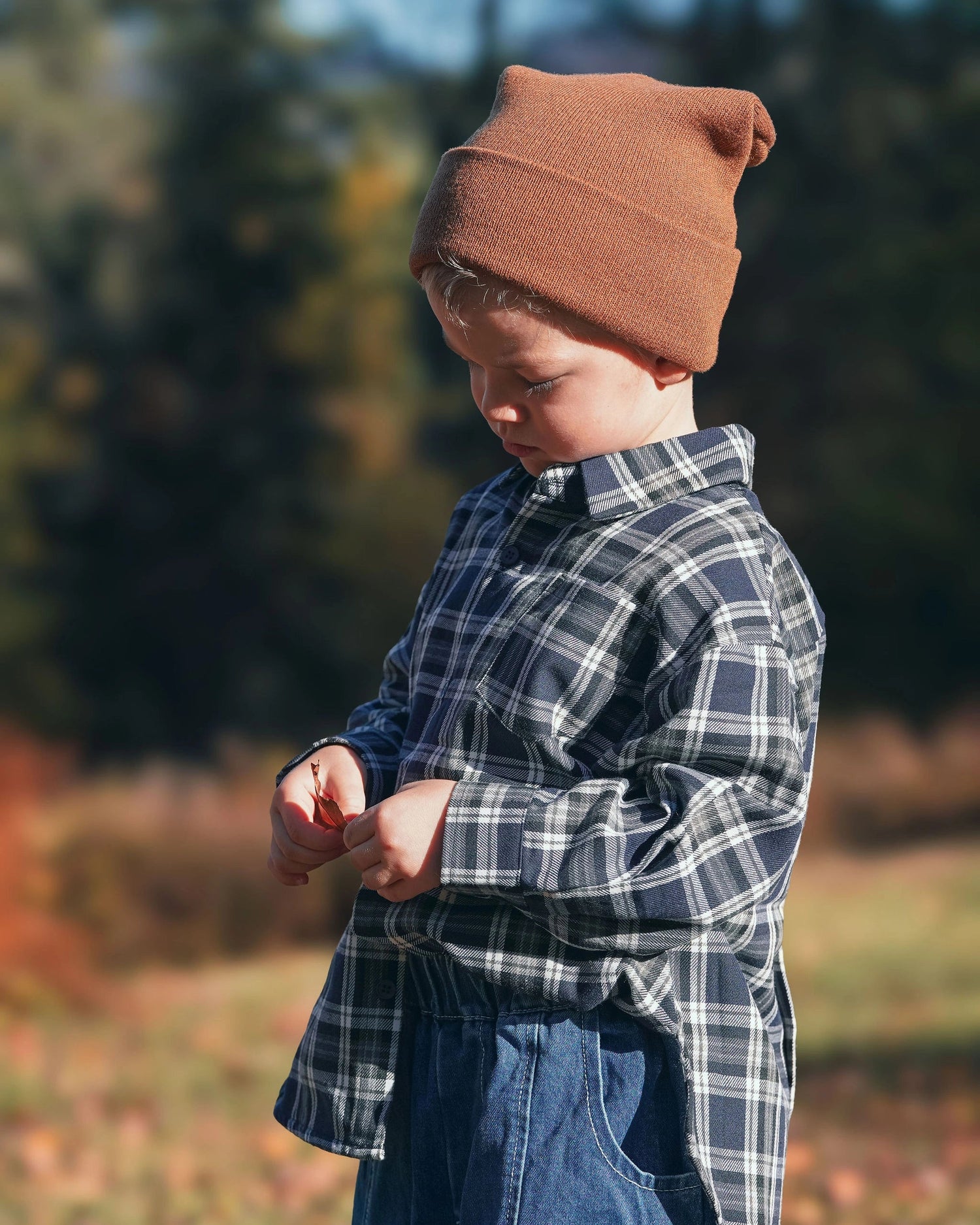 Child wearing a plaid shirt and brown beanie outdoors with trees in the background for Little Aurel